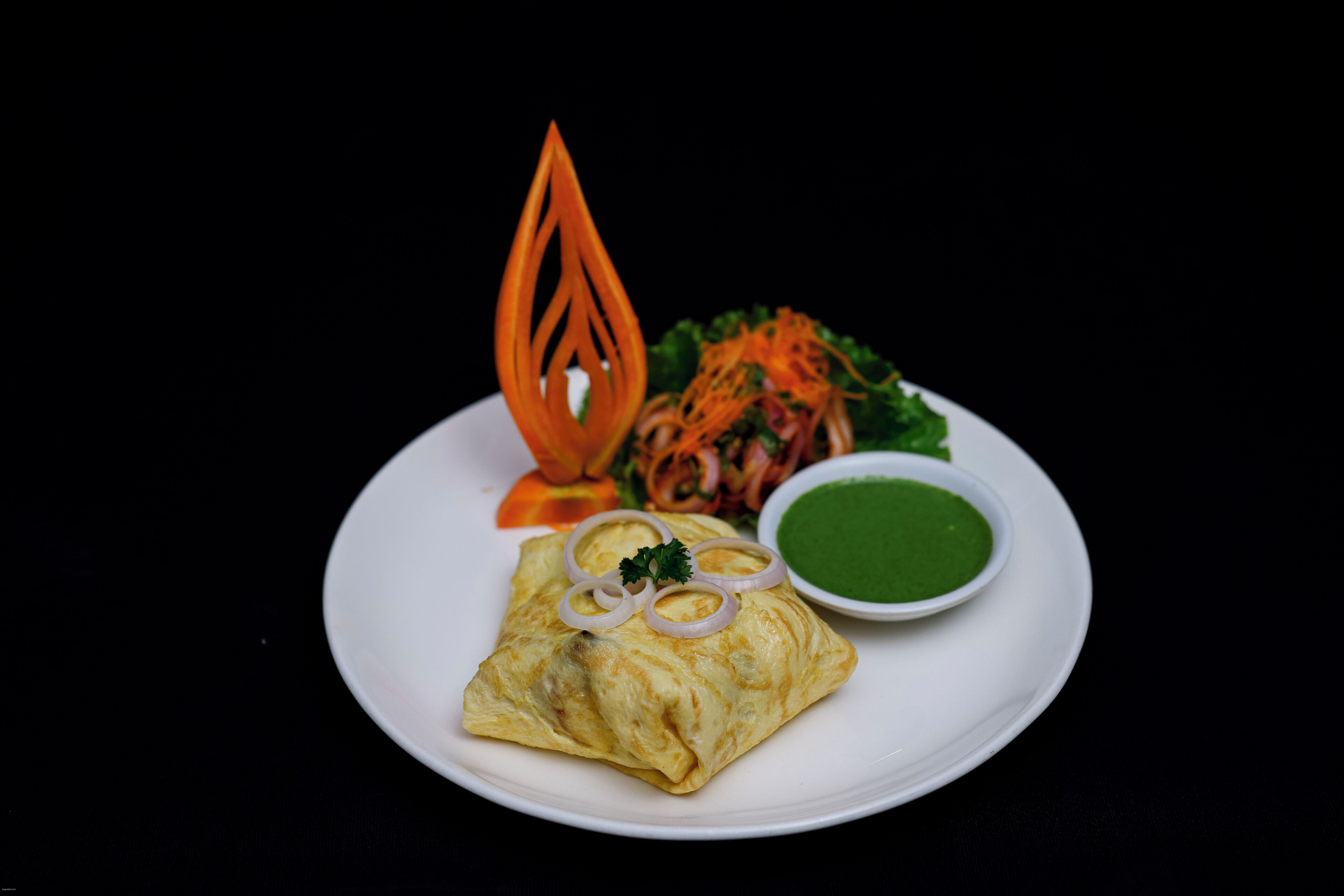 Une assiette contenant une pâtisserie pliée, peut-être une viennoiserie, garnie d’oignons et de persil, accompagnée d’une sauce verte et d’un élément décoratif orange.