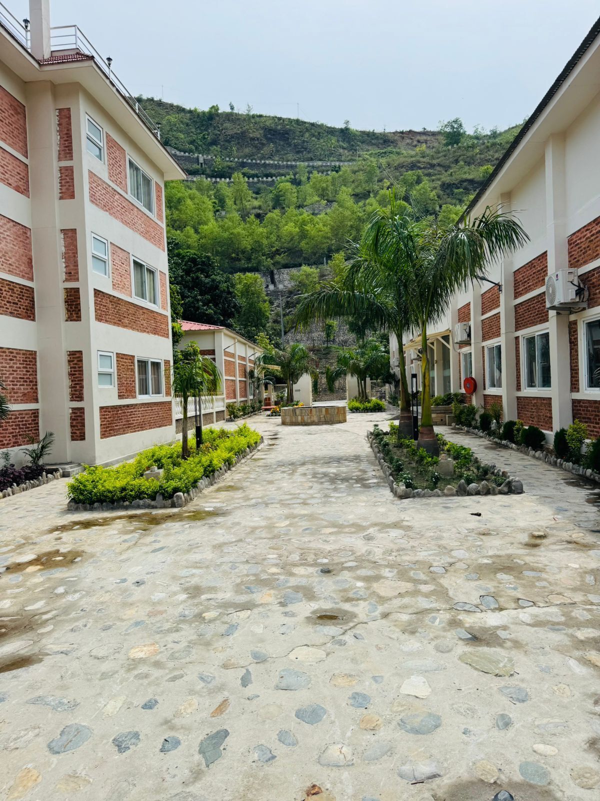 A courtyard with a paved walkway, surrounded by buildings with brick and white facades, and a hillside in the background.