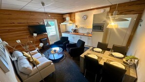 En kompakt opholdsstue med sofa, sofabord, spisebord til fire personer, et køkken med komfur og mikroovn, samt et fjernsyn på væggen.