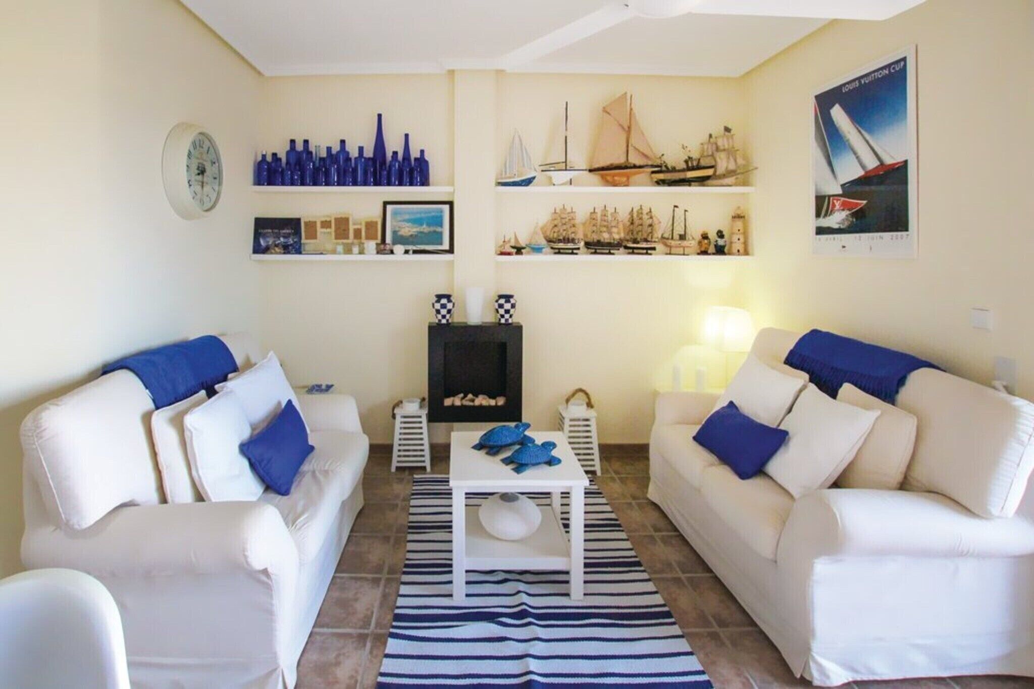 Sala de estar com dois sofás brancos, uma mesa de centro e lareira. Há estantes com objetos decorativos e um cartaz na parede.