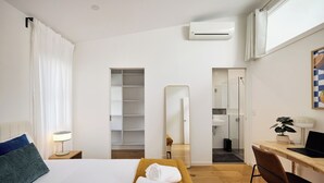 A hotel room with a bed, a desk, a chair, a mirror, a wardrobe, and an air conditioning unit.
