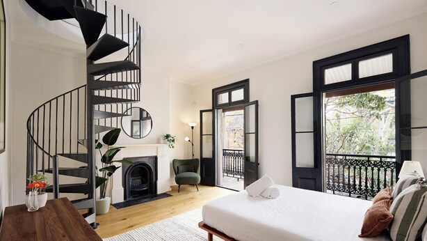 A bedroom with a bed, a wooden nightstand, a black spiral staircase, a fireplace, a green armchair, and a balcony with a black railing.