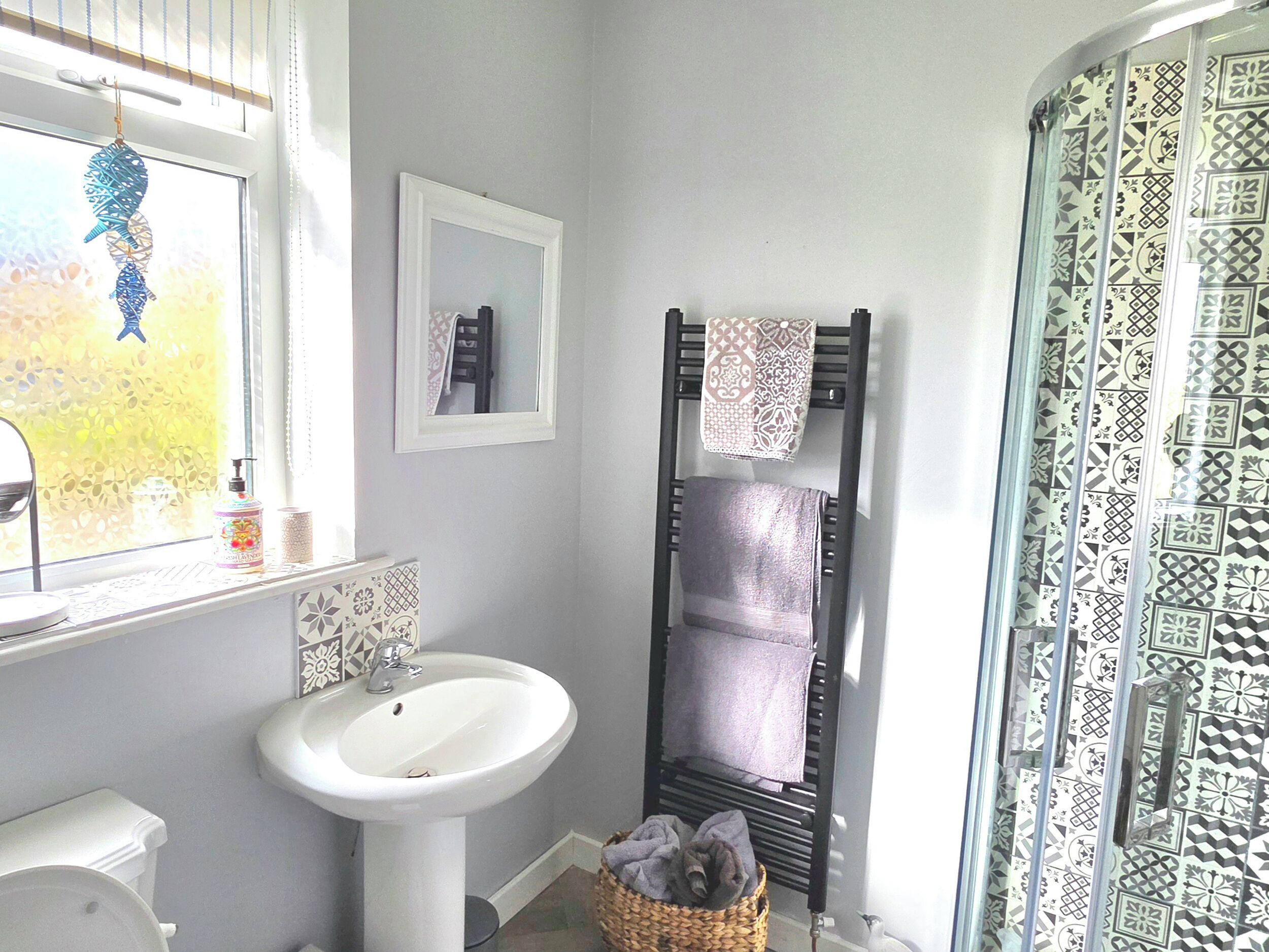 Casa de banho com lavatório branco, espelho, toalheiro com toalha e chuveiro com azulejos estampados.