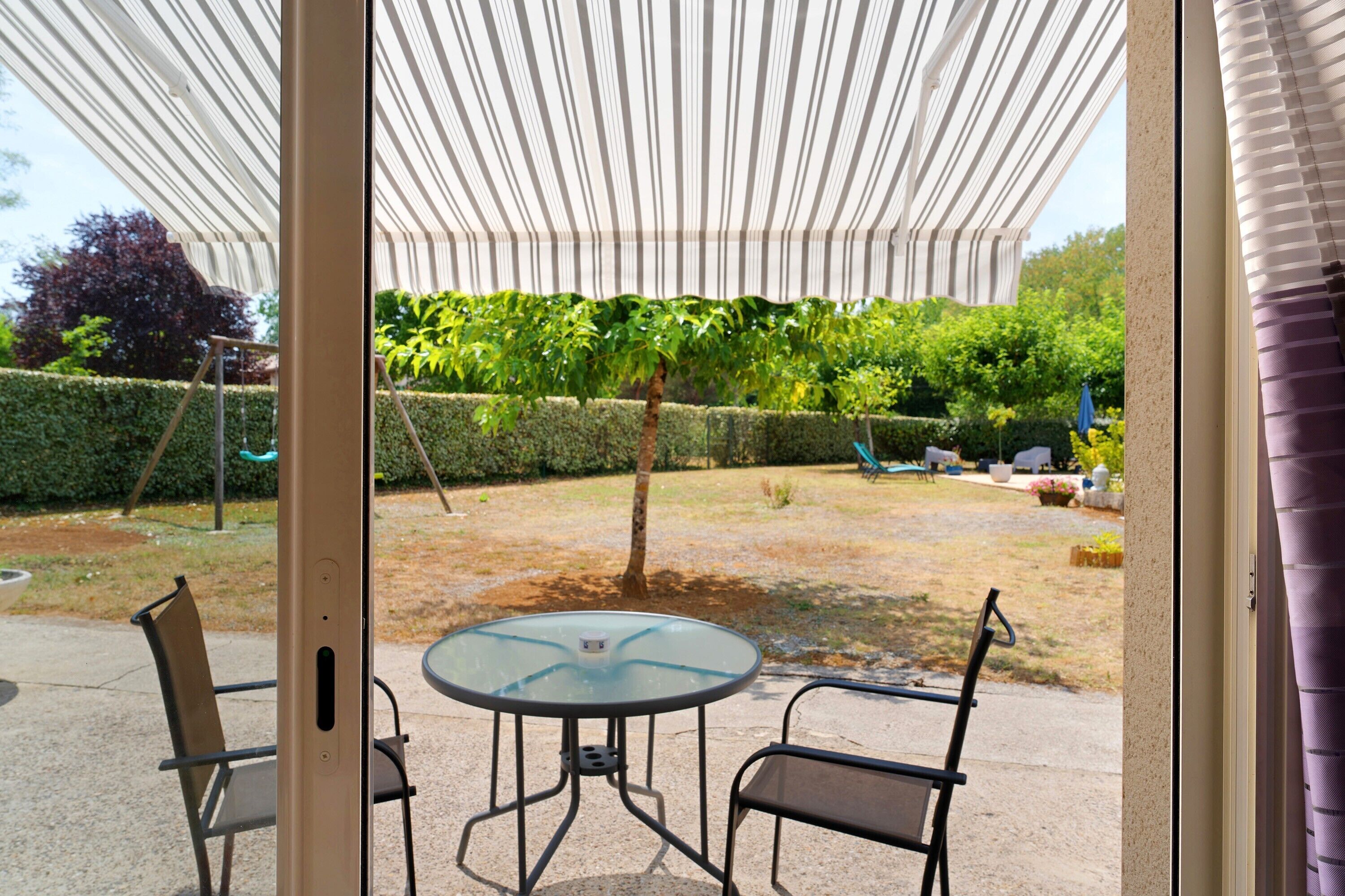 Un patio exterior con mesa y sillas de vidrio, un toldo a rayas y vistas a un jardín ajardinado.