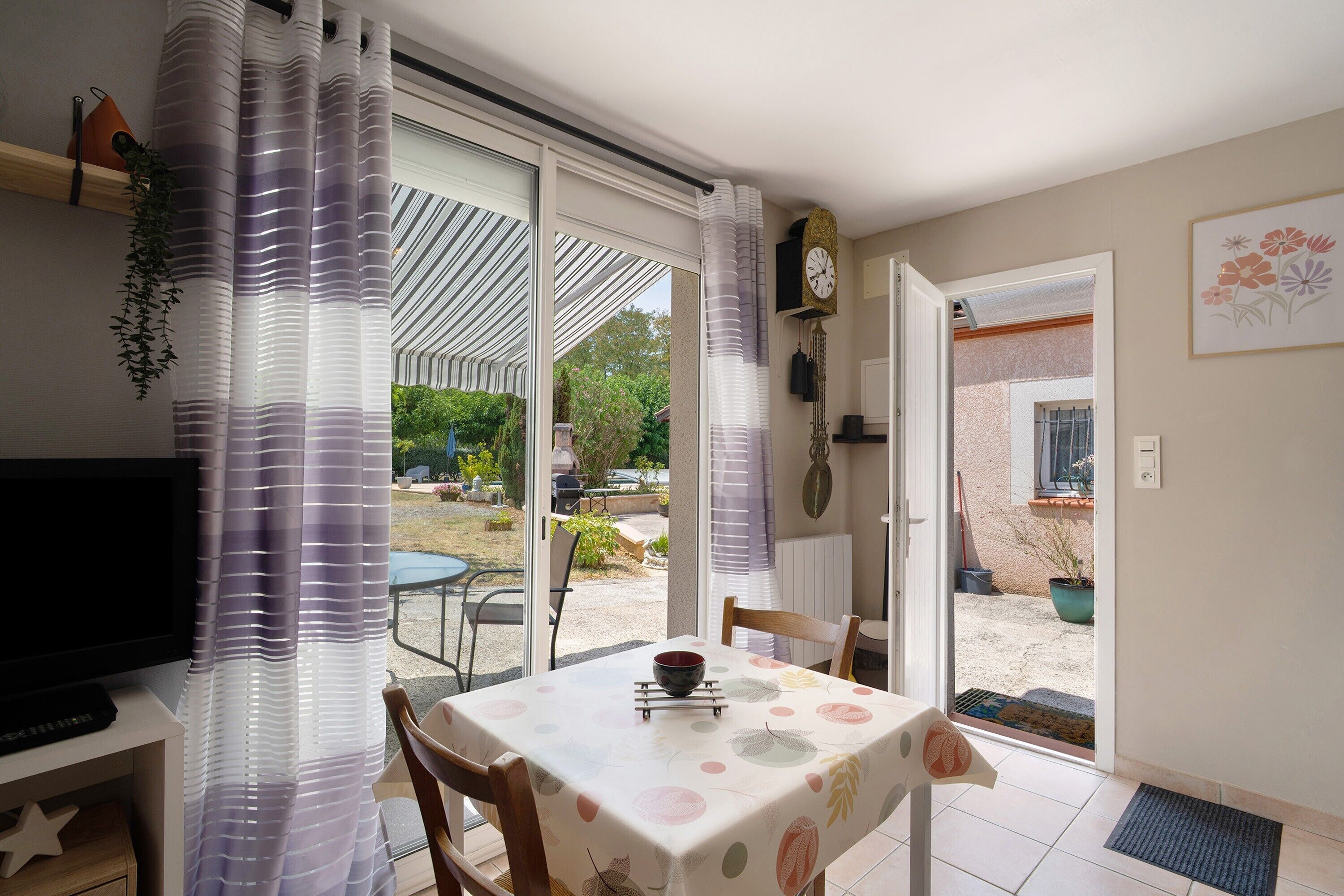 A room with a table, chairs, a TV, a clock, and a view of the outside.