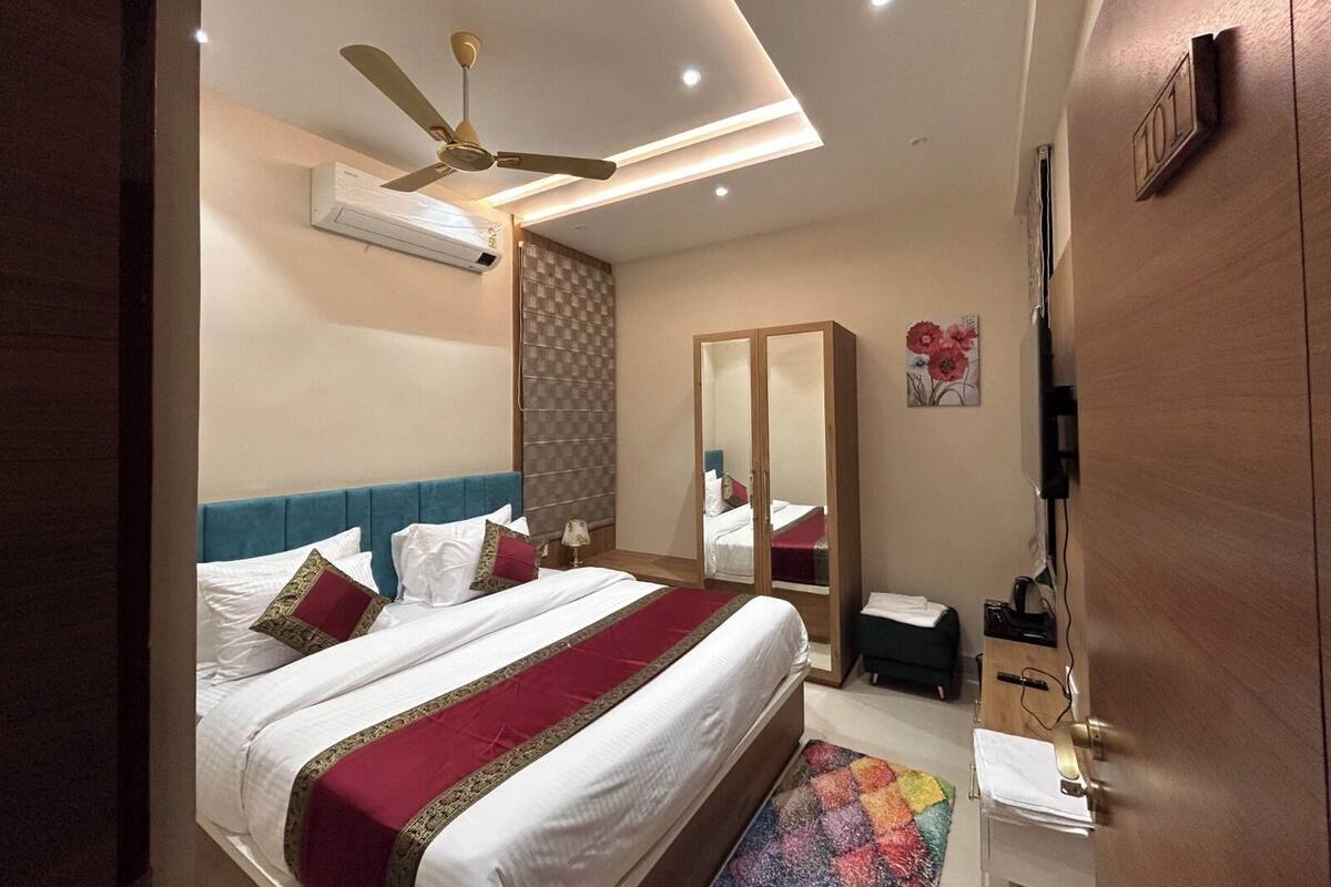 Una habitación de hotel con cama, ventilador de techo, espejo, mesita de noche y una alfombra pequeña.