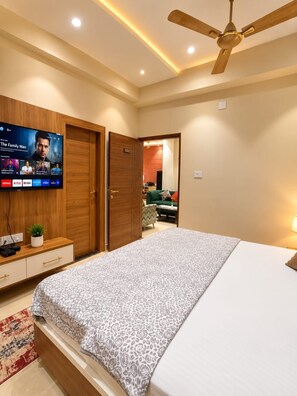 A modern bedroom with a bed, a TV mounted on the wall, a ceiling fan, and a door leading to another room.