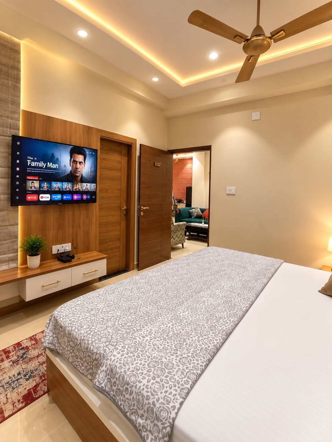 Un dormitorio con cama, televisor montado en la pared, ventilador de techo y puerta que da a otra habitación.