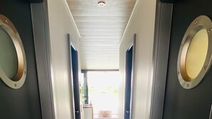 Un couloir moderne aux murs sombres et au plancher en bois clair, qui mène à une pièce lumineuse dotée d’une fenêtre.