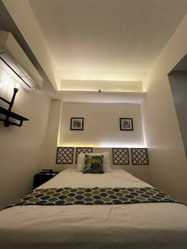 Habitació d'hotel amb un llit, una taula de nit, una estant penjada a la paret i dues fotografies emmarcades.