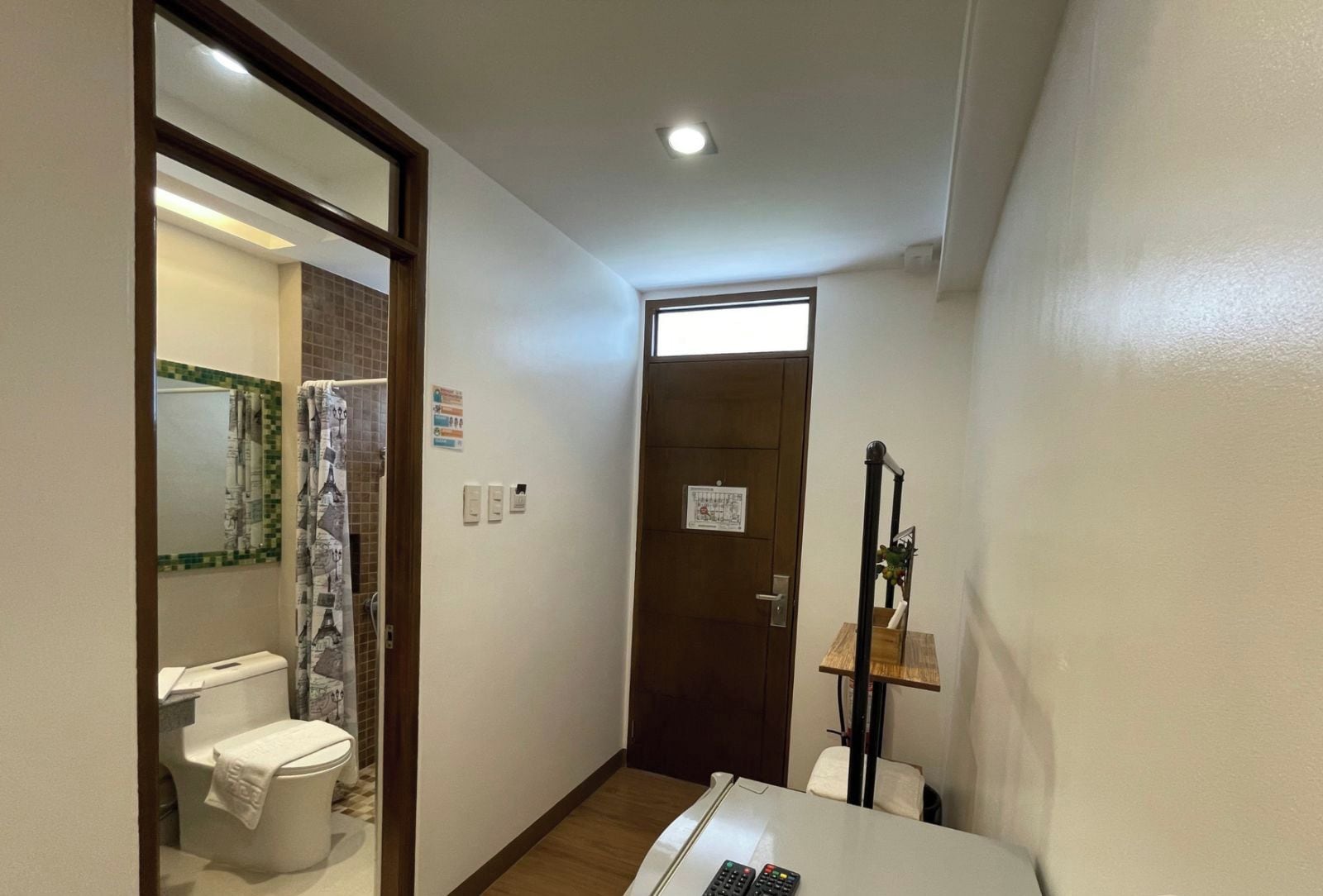 Corredor de hotel com um banheiro, uma mesa pequena com um controle remoto e uma porta que dá para outro quarto.