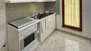 A compact kitchen with white cabinets, a tiled backsplash, a stove, and a window with curtains. - Mistral's House - Apartment in the heart of Alghero (Alghero)