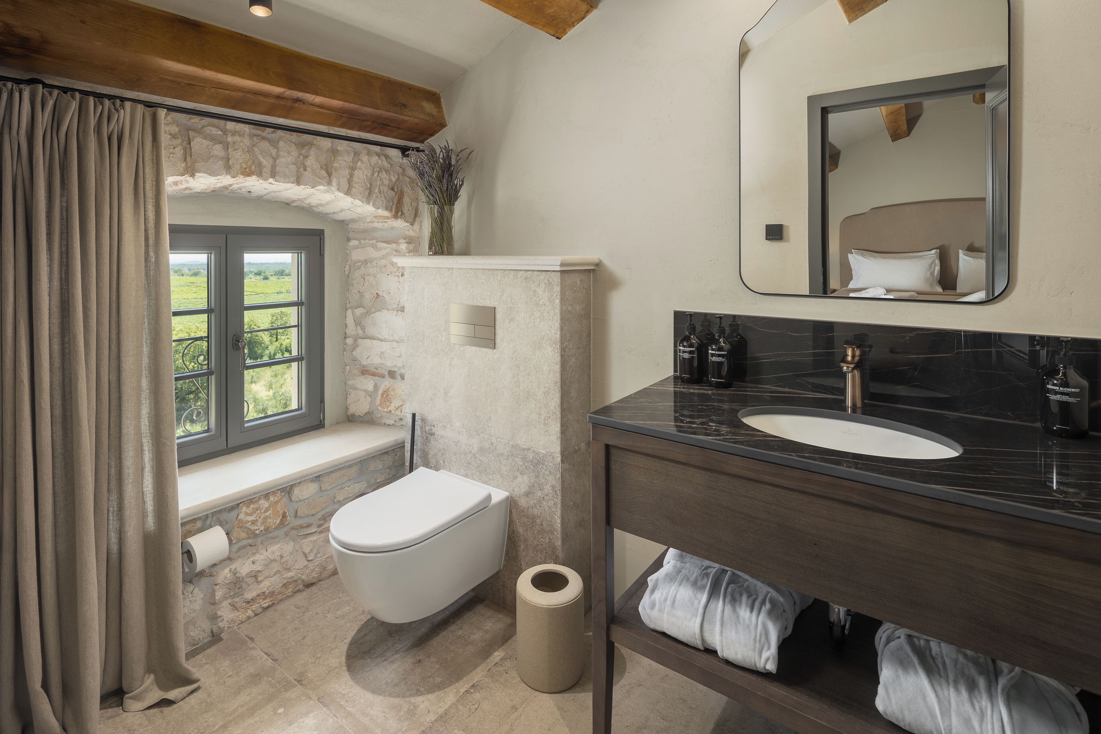 A bathroom with a stone wall, a window with curtains, a toilet, a sink with a mirror, and a towel rack.