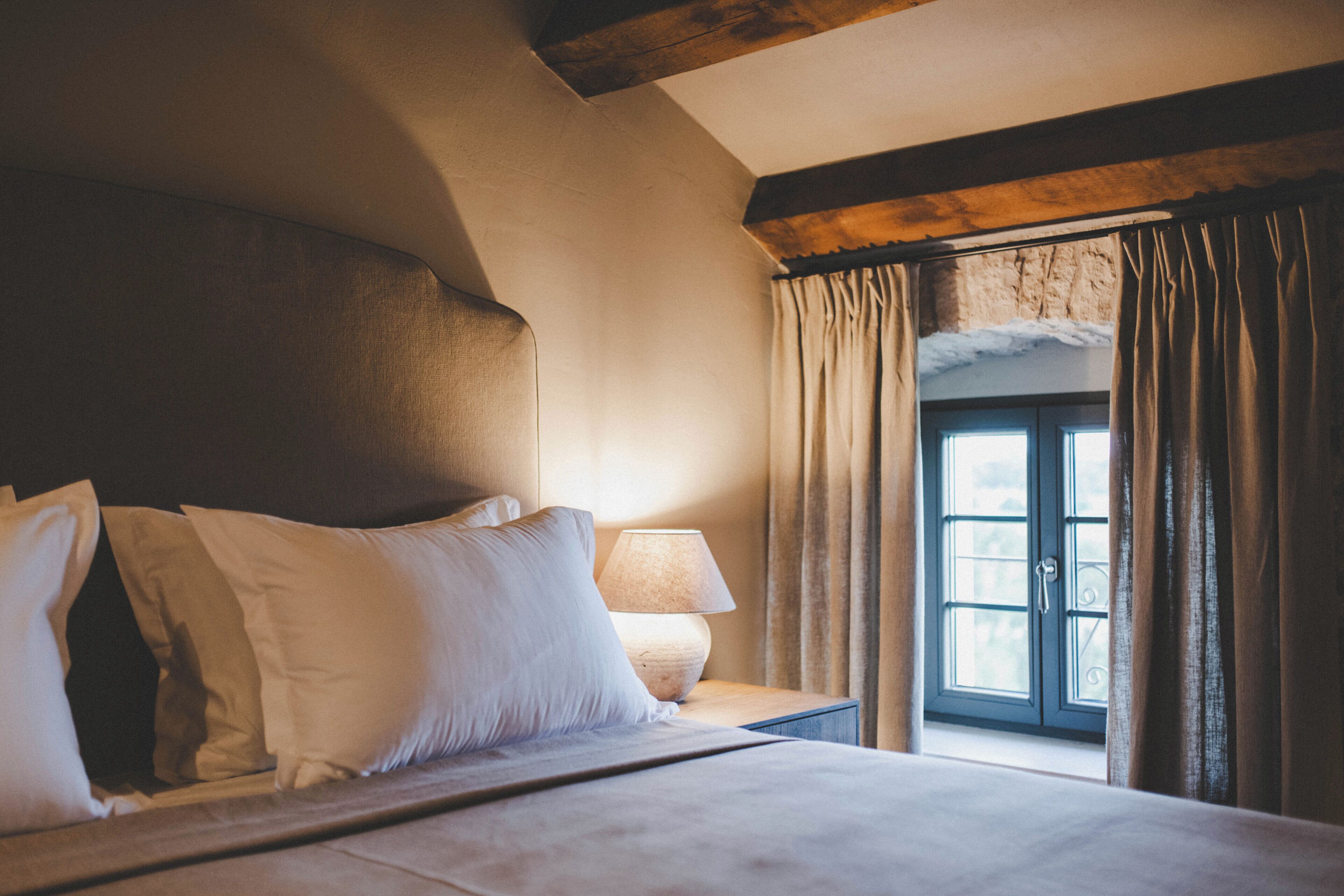 Quarto com cama grande, cabeceira de madeira, mesa de cabeceira com abajur e janela com cortinas.