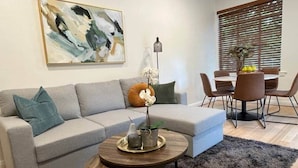 Ein Wohnzimmer mit grauem Sofa, einem runden Couchtisch, einem Gemälde an der Wand und einem Essbereich mit Tisch und Stühlen.