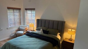 Ein Schlafzimmer mit einem Bett, zwei Nachttischlampen, einem Sessel und zwei Fenstern mit Jalousien.
