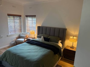Un dormitorio con una cama, dos lámparas de noche, una silla y dos ventanas con persianas.