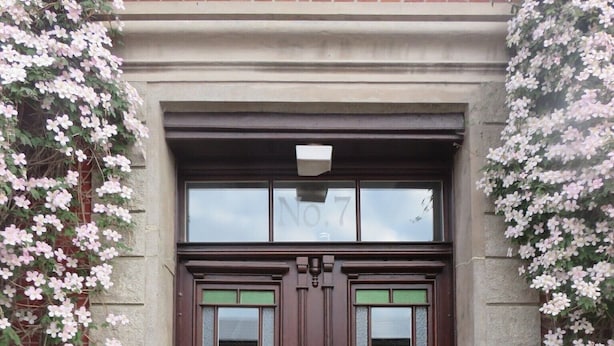 Une porte en bois à deux battants, avec une plaque portant un numéro, une petite marche dotée d’une rampe métallique et un arrangement floral à l’entrée.