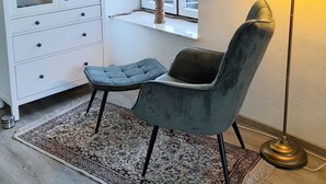 Une pièce avec une bibliothèque blanche, une fenêtre donnant sur des voitures garées, un fauteuil vert, un tapis à motifs et un lampadaire.