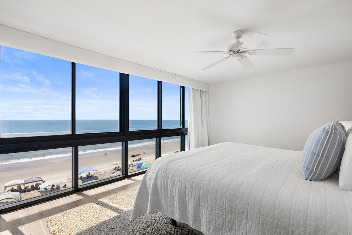 A bedroom with a large window overlooking a beach, a bed with a white bedspread, and a ceiling fan.