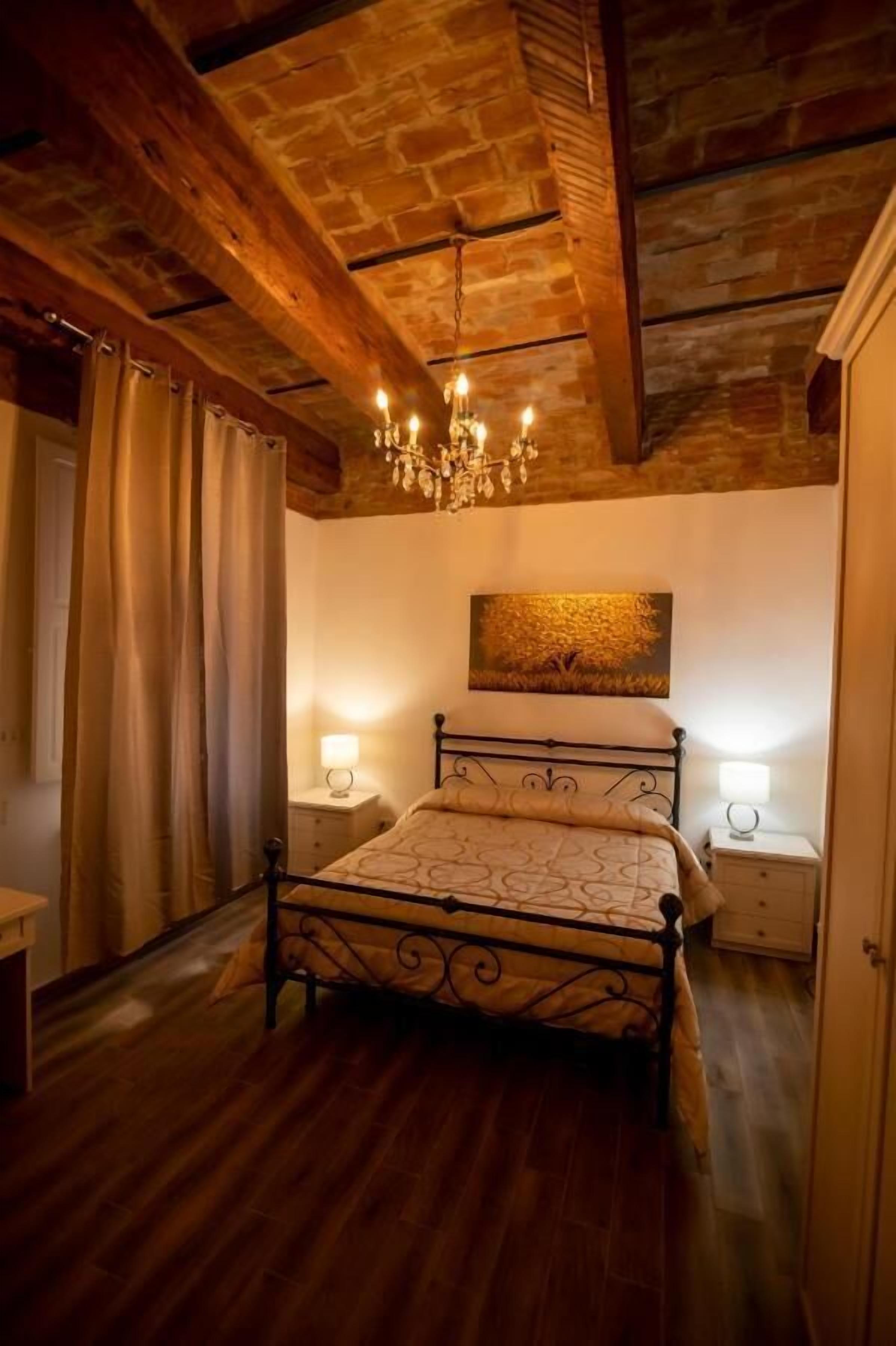 A bedroom with a four-poster bed, wooden beams on the ceiling, a chandelier, and two bedside tables with lamps.