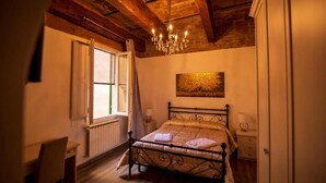 A bedroom with a wooden ceiling, a bed with a gold quilt, a chandelier, a window with curtains, and a painting on the wall. - Le Stanze dei Ghibellini (Florence)