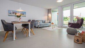 A modern living room with a dining table, grey sofas, a grey pouf, and a large window with a view of greenery. - Beachfront apartment for two people on the pier (Graal-Müritz)