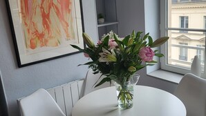 Een moderne kamer met een ronde witte tafel, twee witte stoelen, een vaas met bloemen, een lijst met een abstract schilderij en uitzicht op gebouwen door een raam.
