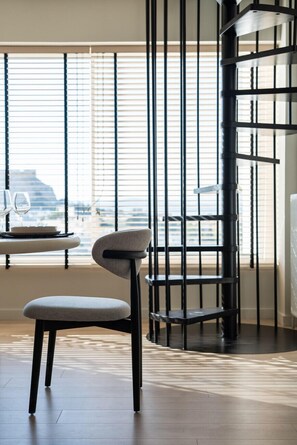 A modern interior with a dining area, a black metal spiral staircase, and large windows with blinds.