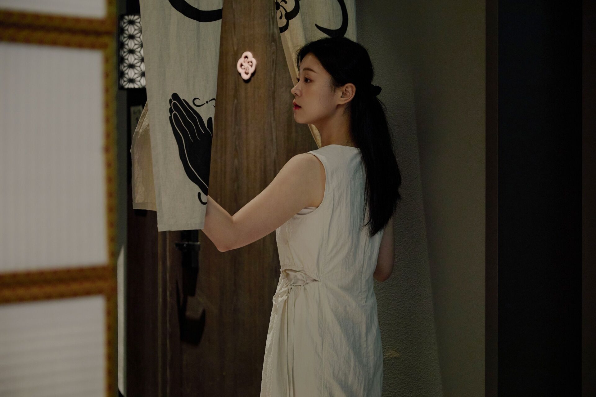 A person in a white dress standing in a room with a wooden door and a curtain with a black design.