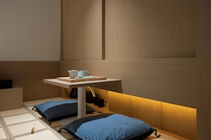 Une petite pièce chaleureuse avec un mur en bois, une table basse sur laquelle se trouvent une bouilloire et des tasses, et deux coussins à motifs posés sur le sol.