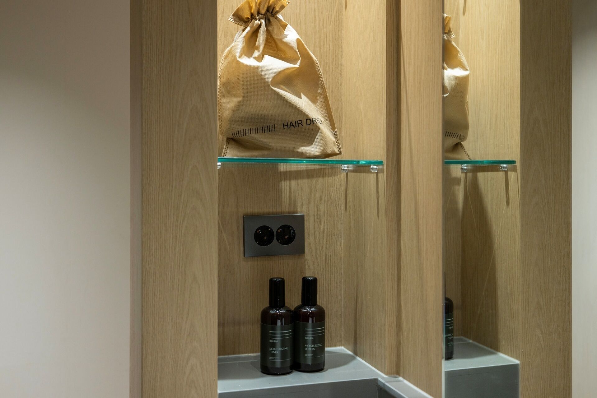 A bathroom with a wooden vanity, a glass shelf holding a hair dryer, and two bottles of moisturizing lotion.