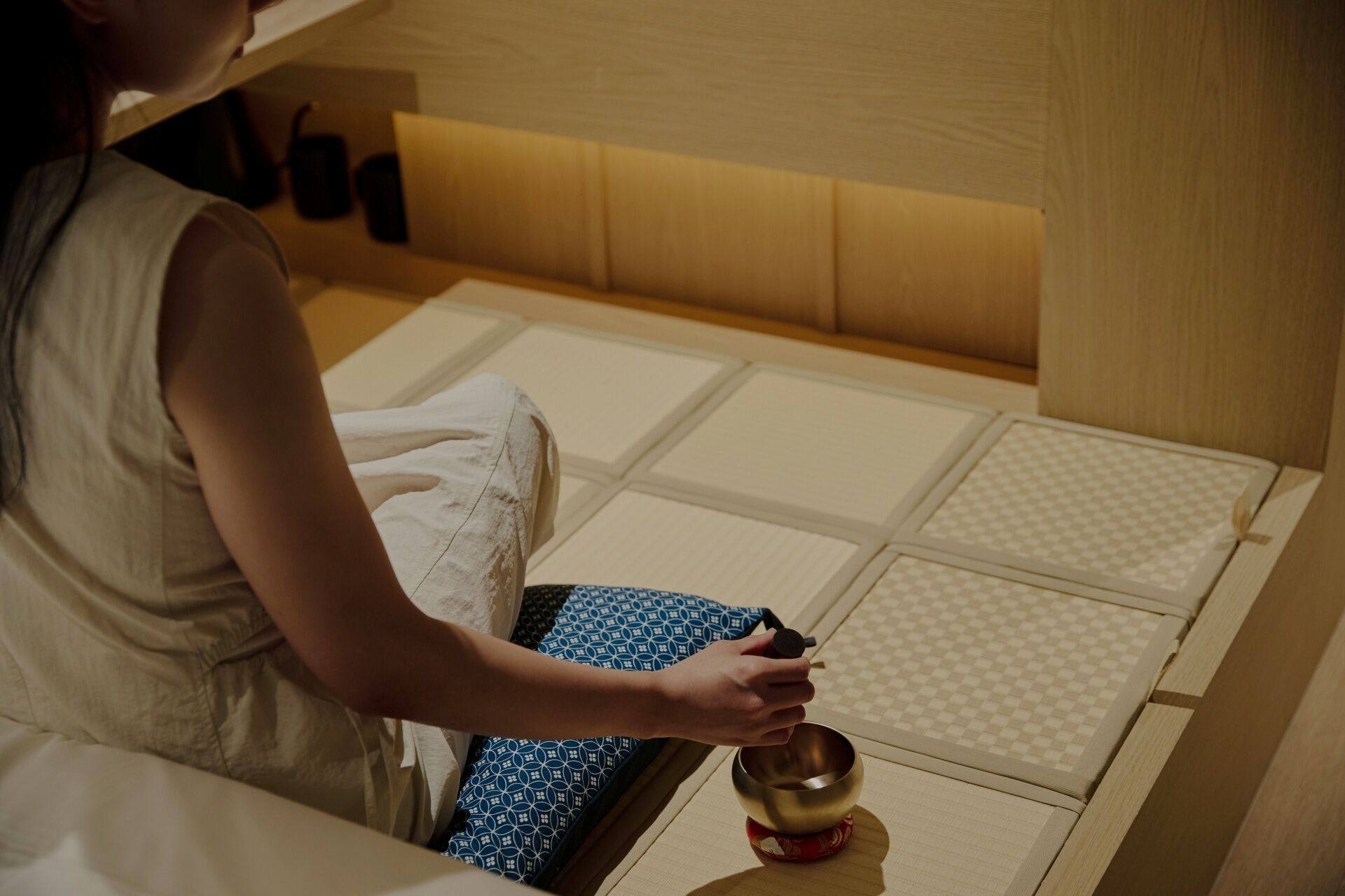 A person sitting on a bed with a patterned cushion, holding a small bowl.