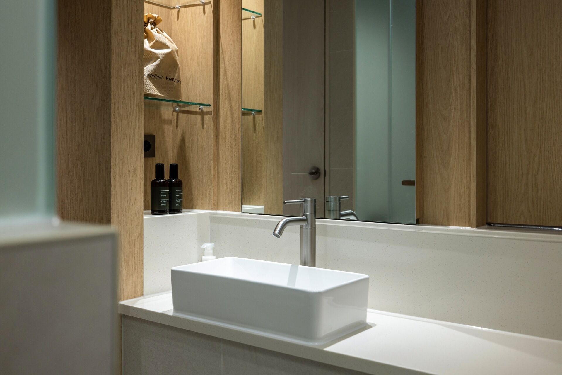 A modern bathroom with a white sink, a glass shelf with toiletries, and a large mirror.