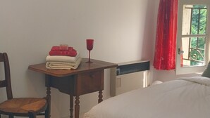 A bedroom with a bed, a wooden chair, a small table with a glass, and a window with red curtains. - Guest Room “La Lagune” with Shared Terrace, Private Garden and Wi-Fi (Saint-André-de-Cubzac)
