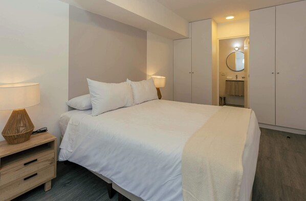 Une chambre à coucher avec un lit, des tables de chevet, une armoire et une salle de bain visible à travers une porte ouverte.