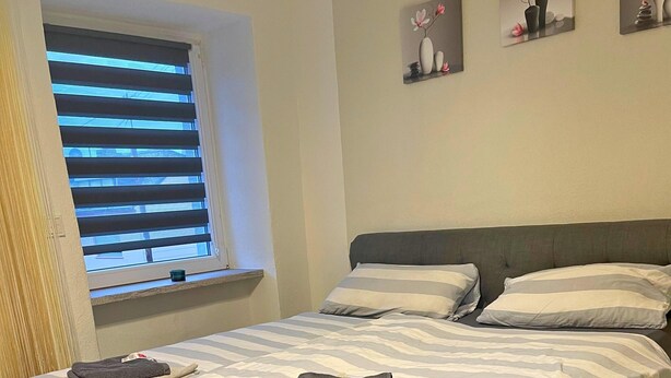 A bedroom with a bed, two pillows, a window with blinds, and three wall-mounted pictures. - Apartment in Trier With Parking (Trier)