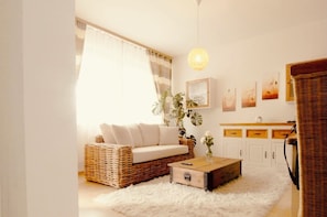 A cozy living room with a wicker sofa, a wooden coffee table, and a white furry rug. There is a plant on the coffee table and a hanging light fixture above.