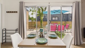 Un coin repas avec une table dressée pour quatre personnes, un étagère en métal et une vue sur le jardin, où se trouve du mobilier d’extérieur.