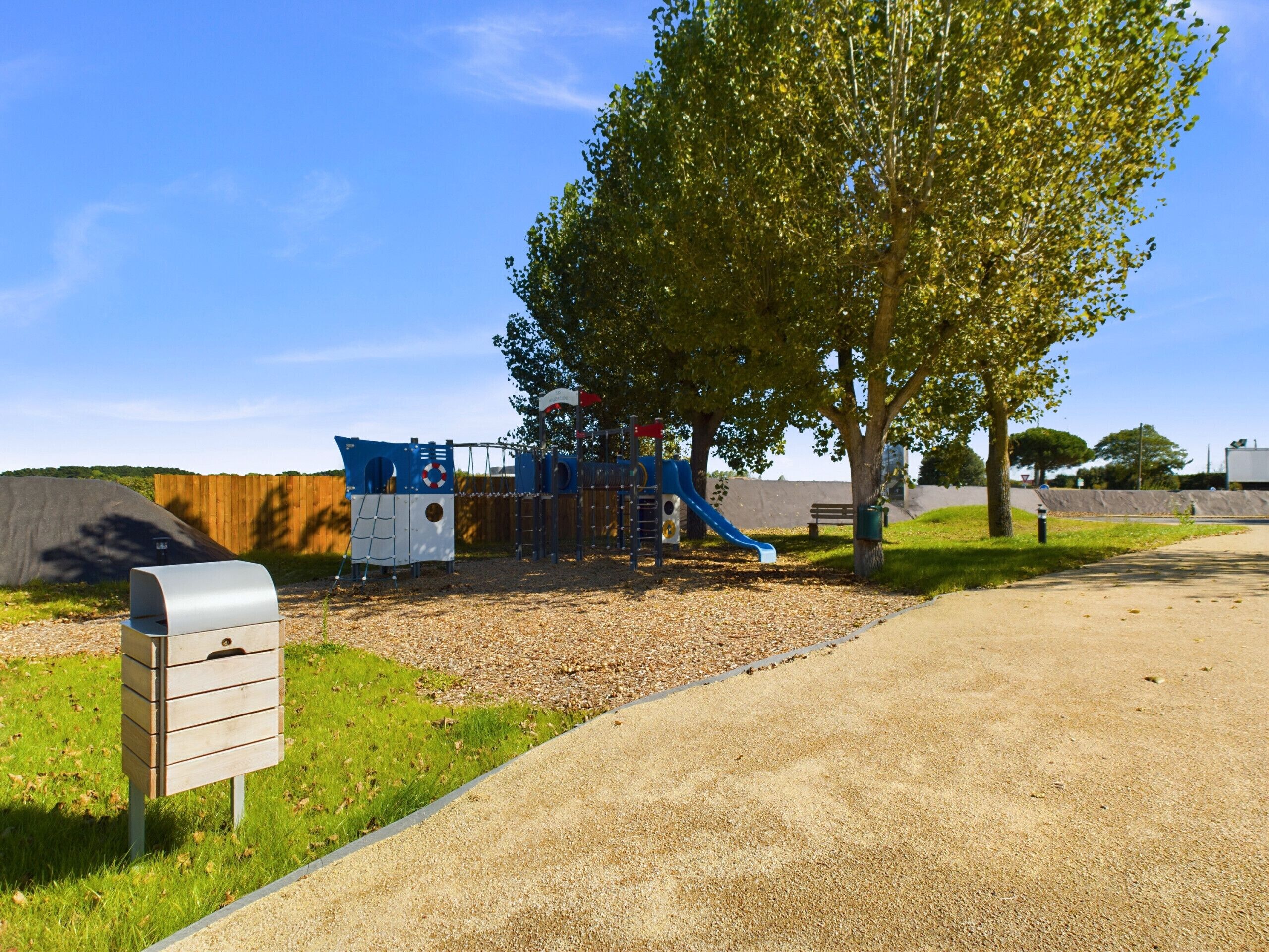A playground with a slide and climbing structures, surrounded by trees and a gravel path.