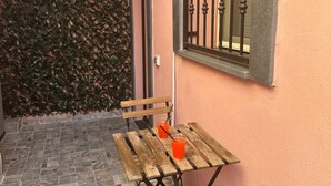 Een terras met een houten tafel en banken, een raam met metalen tralies en een muur begroeid met groene beplanting.