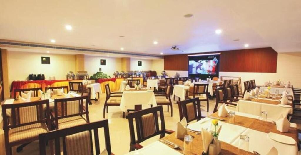 A hotel dining area with multiple tables and chairs, a television screen, and a stage with a red backdrop.