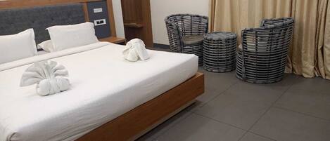 Een netjes opgemaakt bed met witte lakens en handdoeken, een houten hoofdbord, een klein nachtkastje en een rieten stoel.