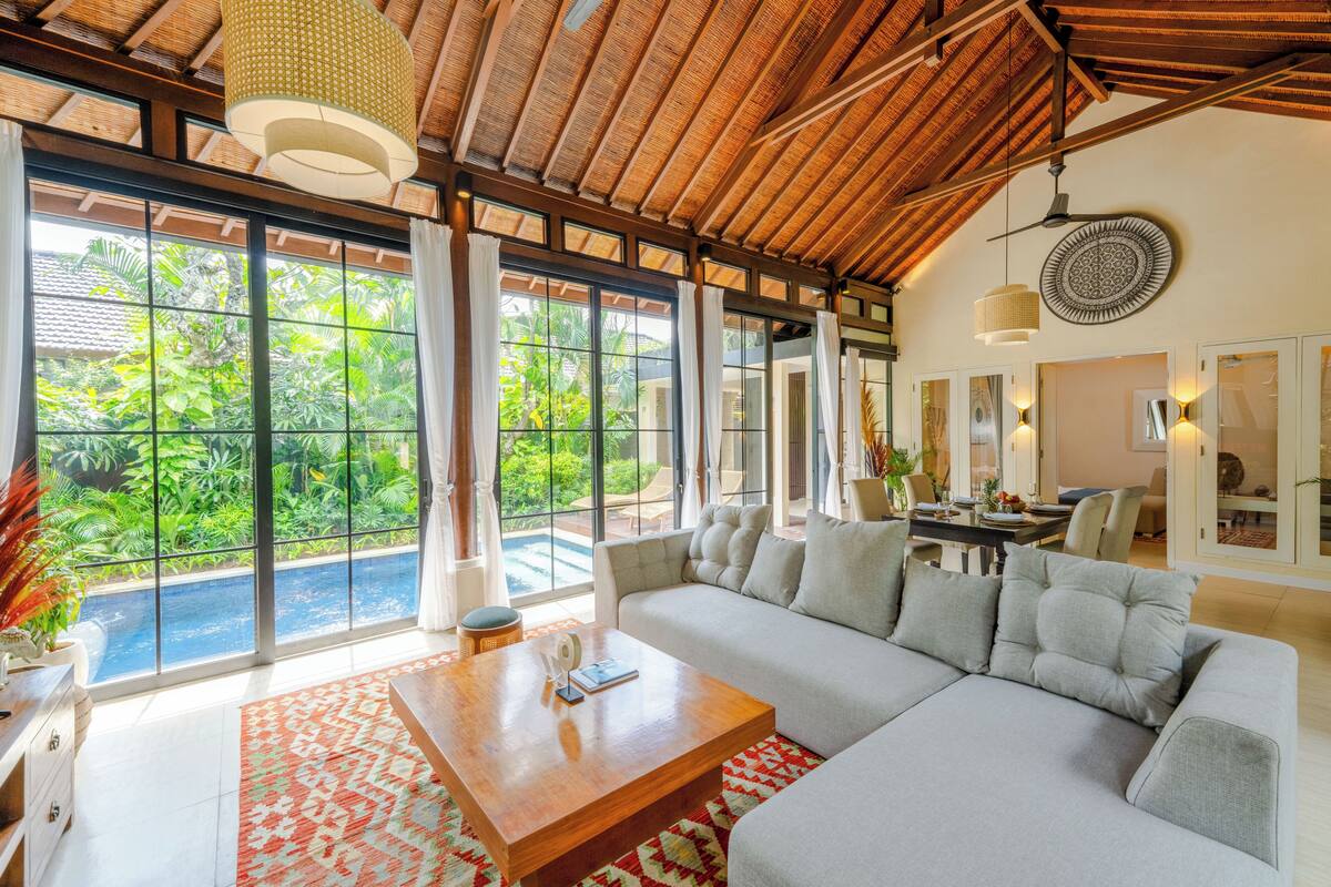 A spacious living area with a large sofa, a wooden coffee table, and a view of a swimming pool and lush greenery.