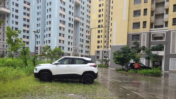 Un VUS blanc est garé sur un trottoir mouillé, dans un quartier résidentiel, avec de grands immeubles en arrière-plan.