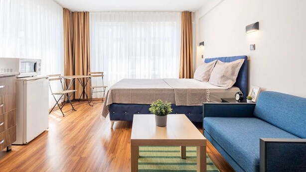 A modern hotel room with a bed, a blue sofa, a small table, a desk with a microwave, and a window with curtains.