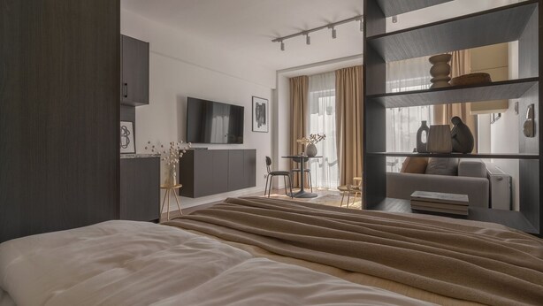 A modern hotel room with a large bed, a TV mounted on the wall, a seating area with a table and chairs, and a bookshelf. - Apartio Rooms Zabrze (Zabrze)