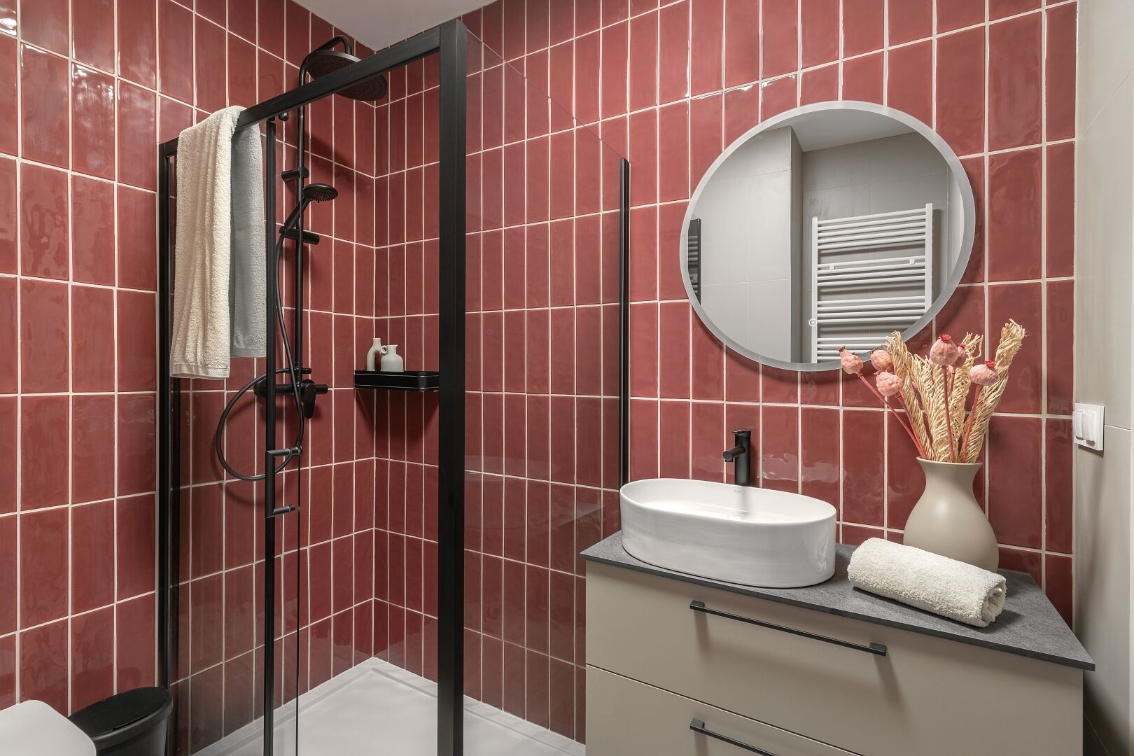 A modern bathroom with a glass-enclosed shower, a round mirror, a white sink, and a vase with dried flowers.