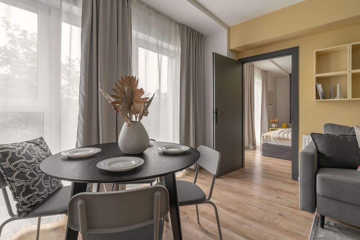 Een moderne woonkamer met een ronde eettafel, twee stoelen, een vaas met gedroogde planten en een grijze bank.