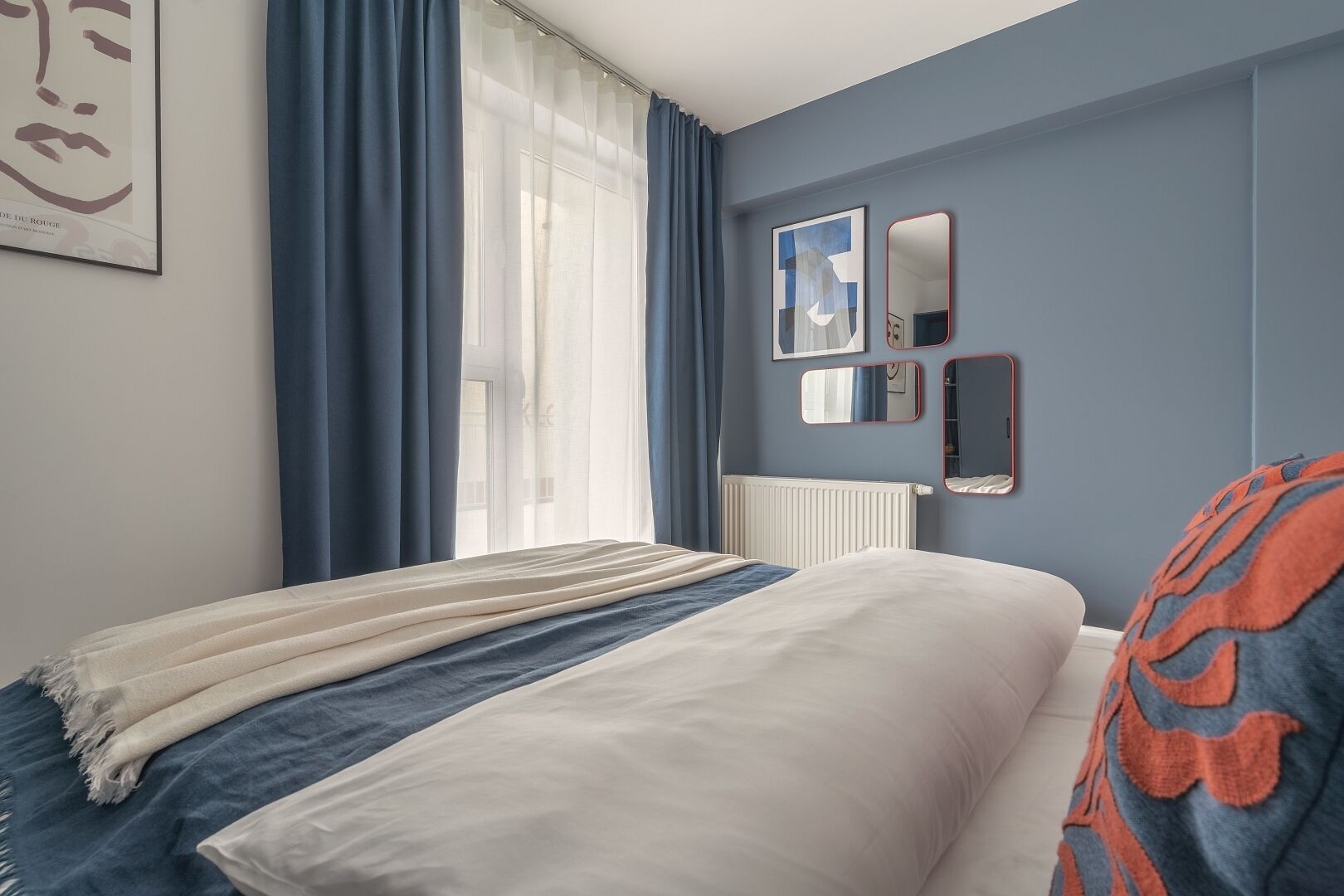 Un dormitorio con cama, ventana con cortinas, radiador y un espejo y obras de arte en la pared.